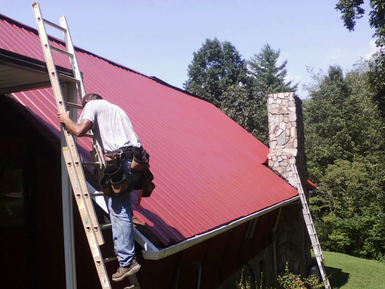Roofer on a ladder beside a red metal roof and stone chimney — Soffit & Fascia Repair by Roofing Brothers in Bretz